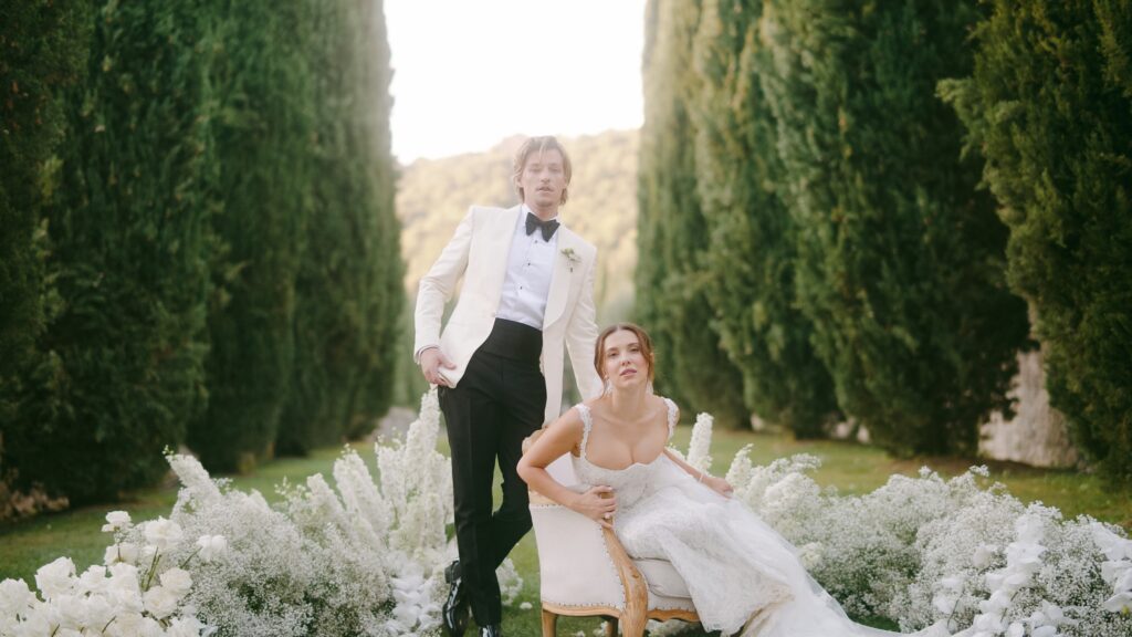 Millie Bobby Brown and Jake Bongiovi standing beneath a floral arch at Villa Cetinale, exchanging vows as the Tuscan sun glows behind them.

