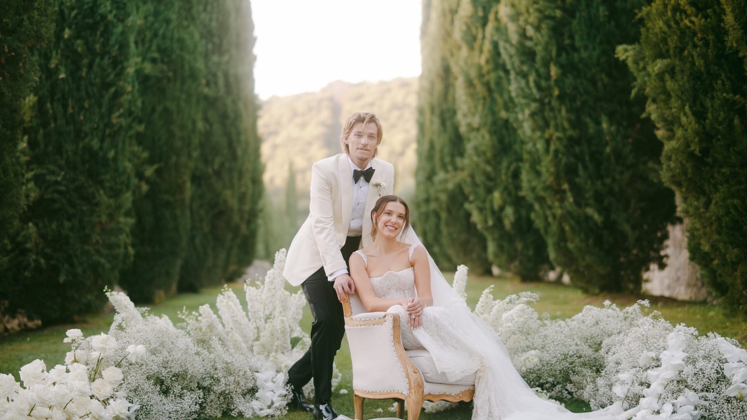 Millie Bobby Brown and Jake Bongiovi standing beneath a floral arch at Villa Cetinale, exchanging vows as the Tuscan sun glows behind them.