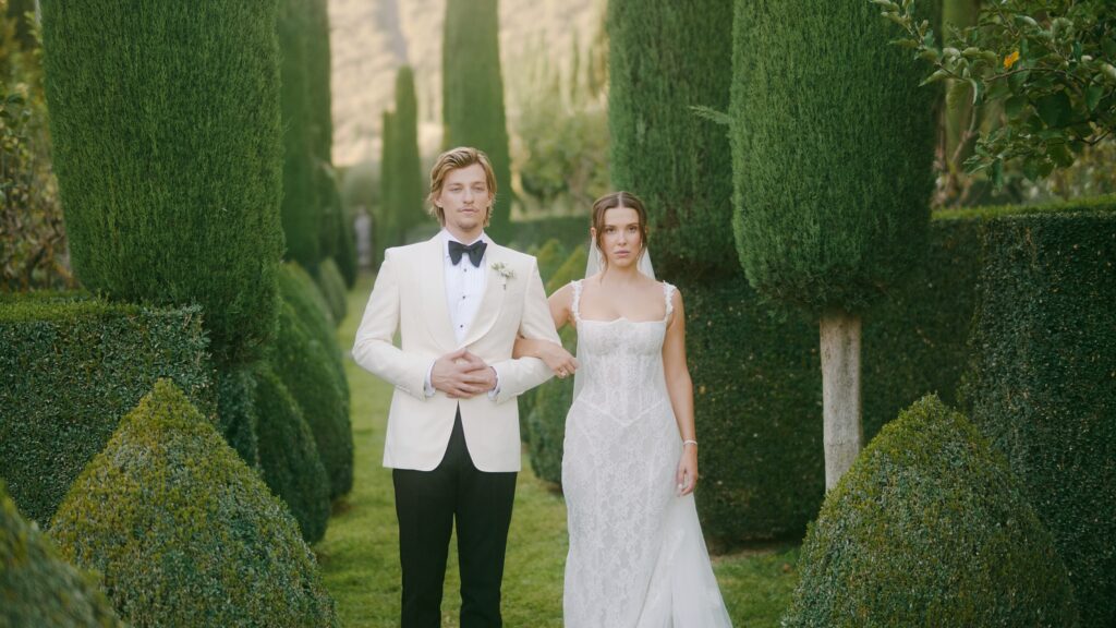 Millie Bobby Brown and Jake Bongiovi standing beneath a floral arch at Villa Cetinale, exchanging vows as the Tuscan sun glows behind them.

