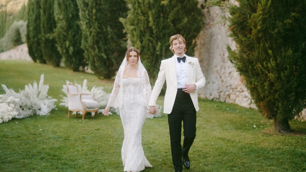 Millie Bobby Brown and Jake Bongiovi standing beneath a floral arch at Villa Cetinale, exchanging vows as the Tuscan sun glows behind them.

