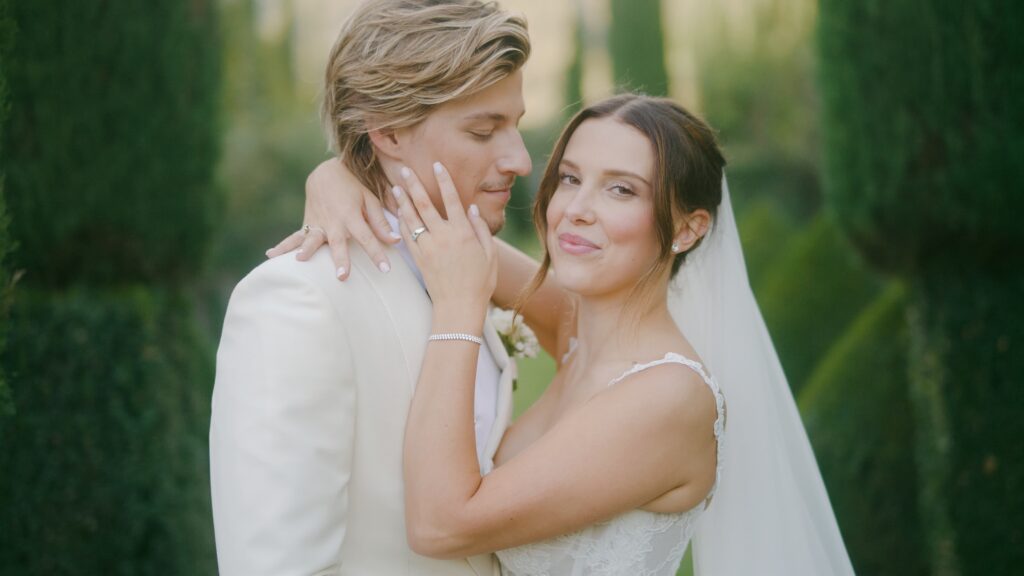 Millie Bobby Brown and Jake Bongiovi standing beneath a floral arch at Villa Cetinale, exchanging vows as the Tuscan sun glows behind them.

