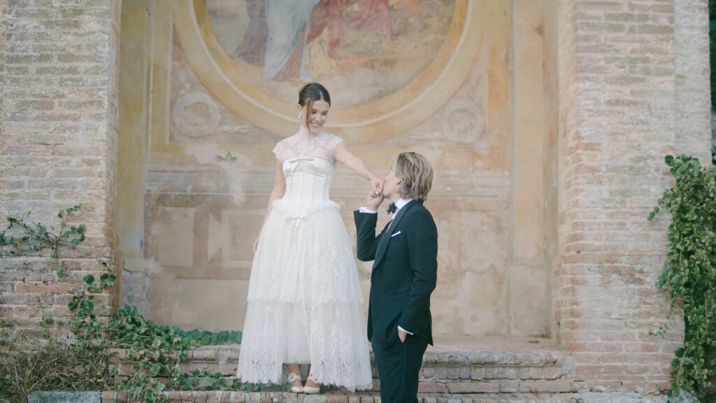 Millie Bobby Brown and Jake Bongiovi standing beneath a floral arch at Villa Cetinale, exchanging vows as the Tuscan sun glows behind them.

